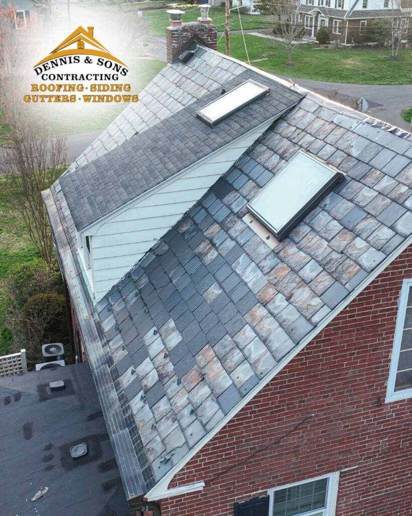 An aerial, high-angle shot captures the multi-level roof of a residential house, highlighting extensive roof repairs. The dominant roof section, constructed from aged grey slate tiles, shows numerous mismatched, lighter grey and tan replacement tiles scattered across its surface, particularly along the lower section and near a large rectangular skylight. A narrower, higher roof section with a smaller skylight runs parallel to it, displaying more uniform grey tiles. To the left, a white dormer wall separates the roof sections, and a brick chimney rises near the top peak. In the lower-left corner, a lower flat roof area features two dark, circular vents. In the upper-left corner, a semi-transparent corporate logo overlay reads, "DENNIS & SONS CONTRACTING ROOFING • SIDING GUTTERS • WINDOWS" in stylized black, white, and gold font, accompanied by a small graphic of a house with a sunburst behind it. The house itself features red brick walls and white-trimmed windows, with a glimpse of a green lawn, trees, a neighboring house, and an asphalt road visible in the blurred background. - dennisandsonscontracting.com